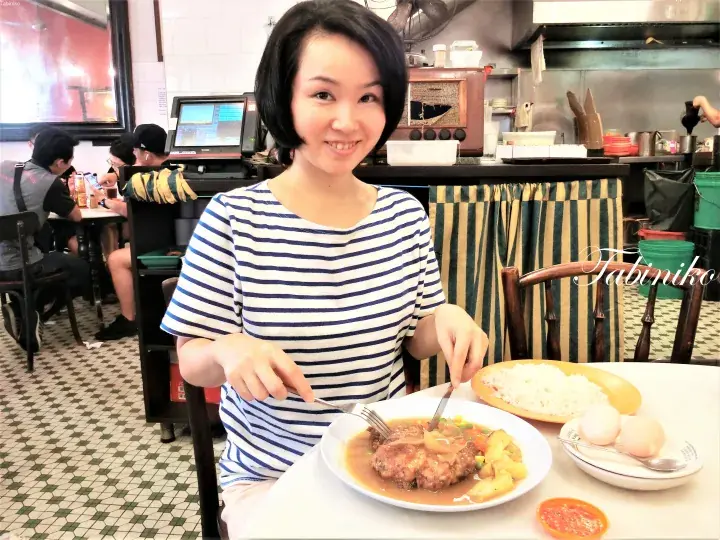 Smiling woman eating a meat patty with gravy and rice at a Malaysian kopitiam.
