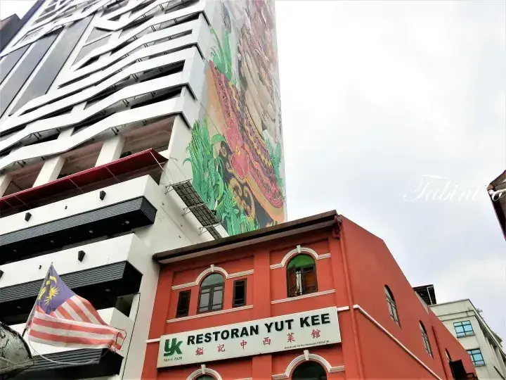 带有大型壁画的现代建筑前,挂着马来西亚国旗的红色“Restoran Yut Kee”餐厅外观。