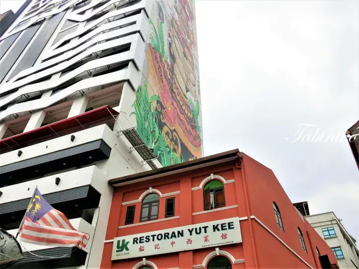 The red facade of Restoran Yut Kee with its sign and a Malaysian flag, set against a modern building with a large mural.