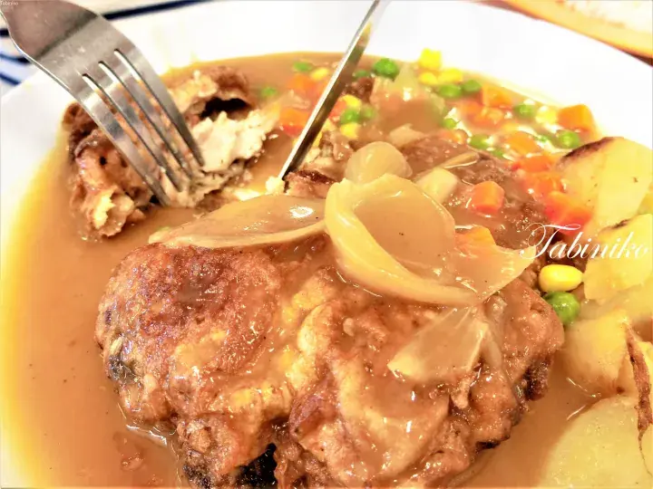 Close-up of a savory meat dish with gravy, onions, and mixed vegetables being cut with a fork and knife.