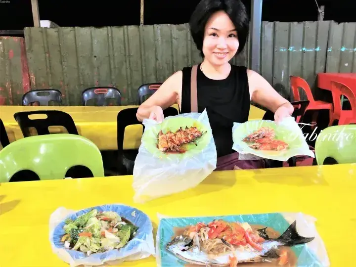 一名女子在户外大排档的黄色桌子旁，手持盛有烤海鲜的盘子微笑。