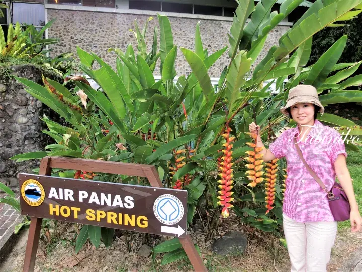 A woman stands beside a sign for Air Panas Hot Spring surrounded by tropical plants.
