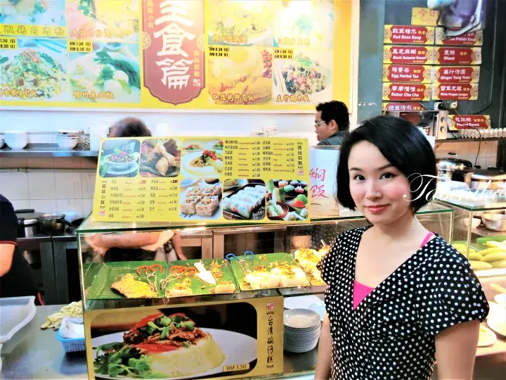 様々なマレーシア料理とメニューが並ぶフードスタールのショーケースの前に立つ女性。