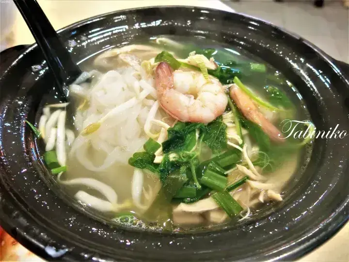 A steaming bowl of clear soup with rice noodles, shrimp, bean sprouts, and greens in a black ceramic pot.