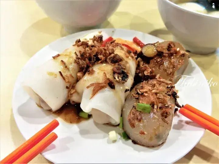 Steamed rice rolls and tau kua kueh topped with fried shallots and sauce, served with chopsticks on a white plate.