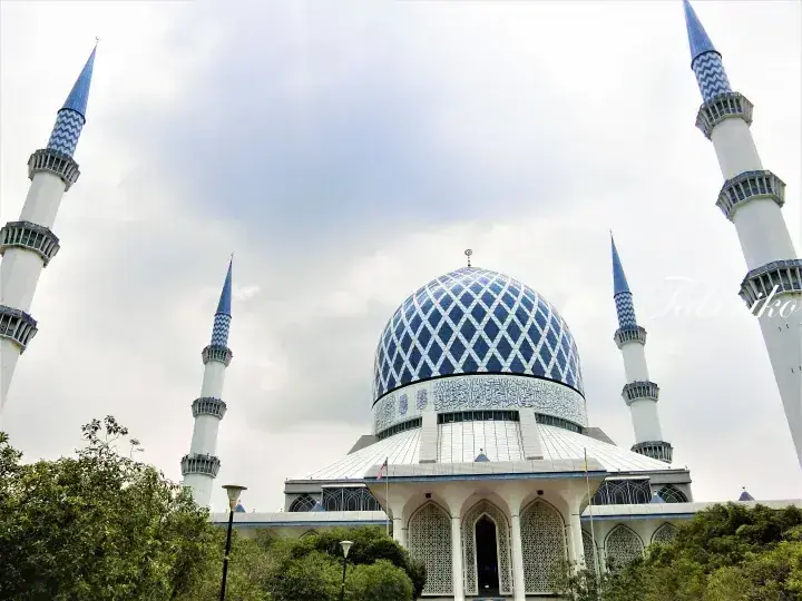 The Sultan Salahuddin Abdul Aziz Shah Mosque featuring its iconic blue dome and four tall minarets against a cloudy sky.