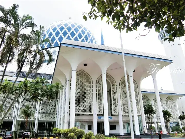The blue dome and white arched entrance of the Sultan Salahuddin Abdul Aziz Shah Mosque surrounded by palm trees.