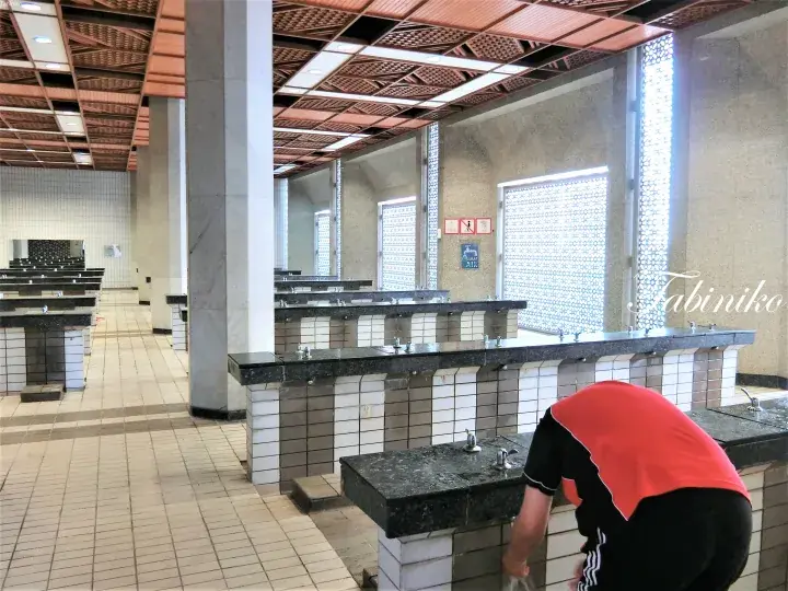 タイル張りの壁と装飾的な天井が特徴的な広々とした公共トイレで、オレンジ色のシャツを着た人物が洗面台を掃除している。
