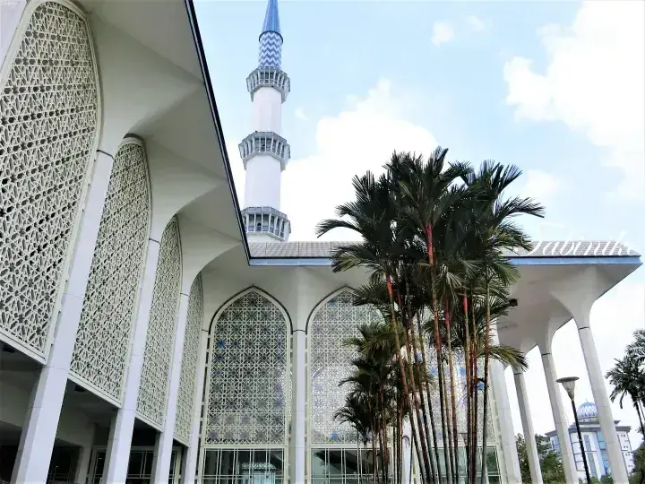 Modern white mosque with intricate lattice patterns and a tall minaret, surrounded by palm trees under a blue sky.