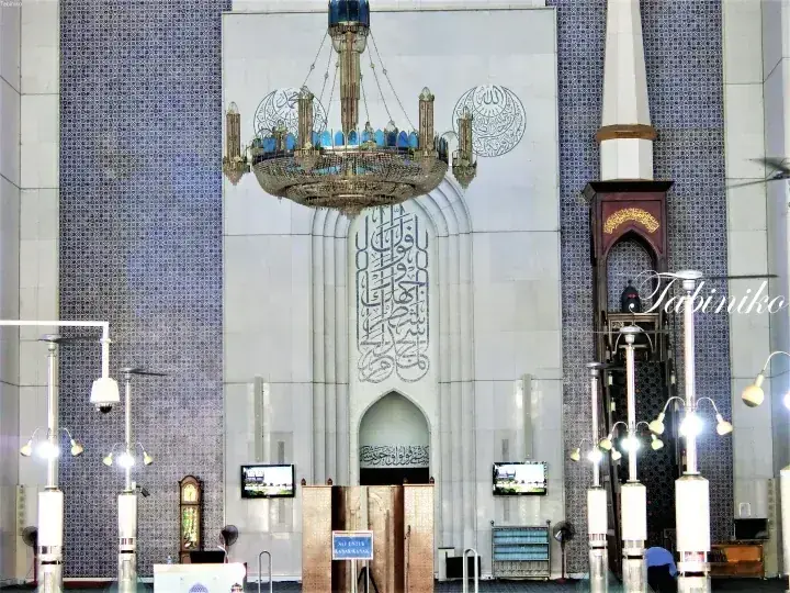 Interior view of a grand mosque featuring a large ornate chandelier, intricate Arabic calligraphy on the wall, and blue mosaic tiles.
