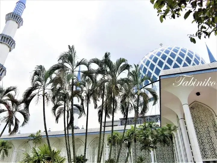 Blue dome and minaret of the Blue Mosque in Shah Alam framed by tall palm trees against a cloudy sky.