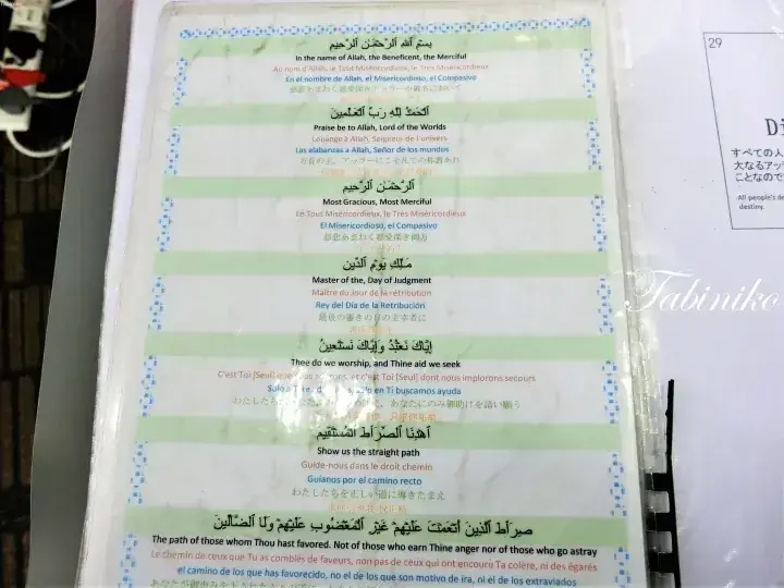 Close-up of Surah Al-Fatiha printed in Arabic with English, French, Spanish, and Japanese translations on a laminated page.