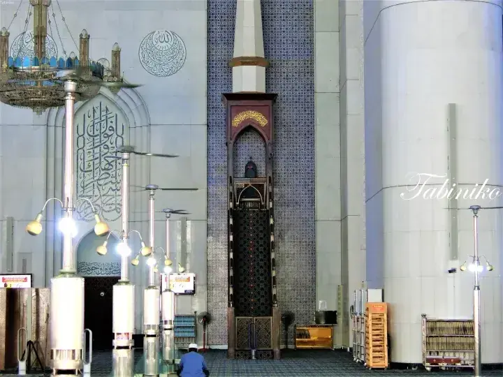 Interior view of a mosque featuring ornate calligraphy, a tall minbar, and decorative lighting fixtures.