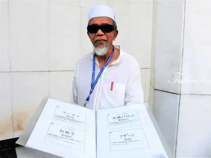 Man in white cap and sunglasses holding a brochure with Japanese text about the Quran and prayer.