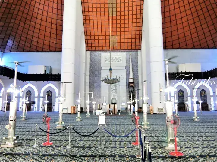 Interior of a grand mosque with tall white pillars, a patterned carpet, and a distinctive orange-tiled ceiling.