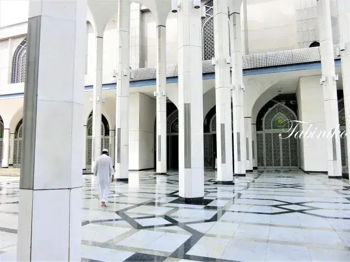 A man in white walks through the modern white pillars and geometric tiled floor of a mosque courtyard.