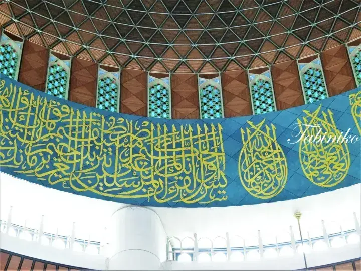 Interior view of a mosque featuring a large blue banner with yellow Arabic calligraphy hanging beneath a geometric dome ceiling.