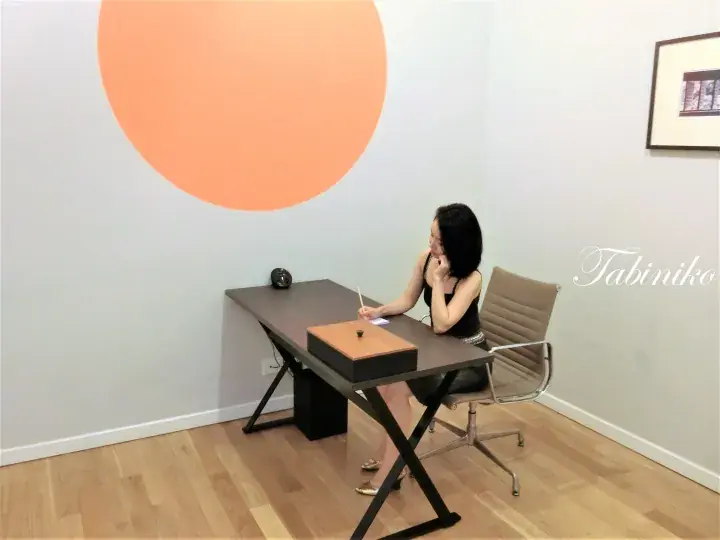 Woman writing at a desk in a modern room with a large orange circle wall art and Tabiniko watermark.