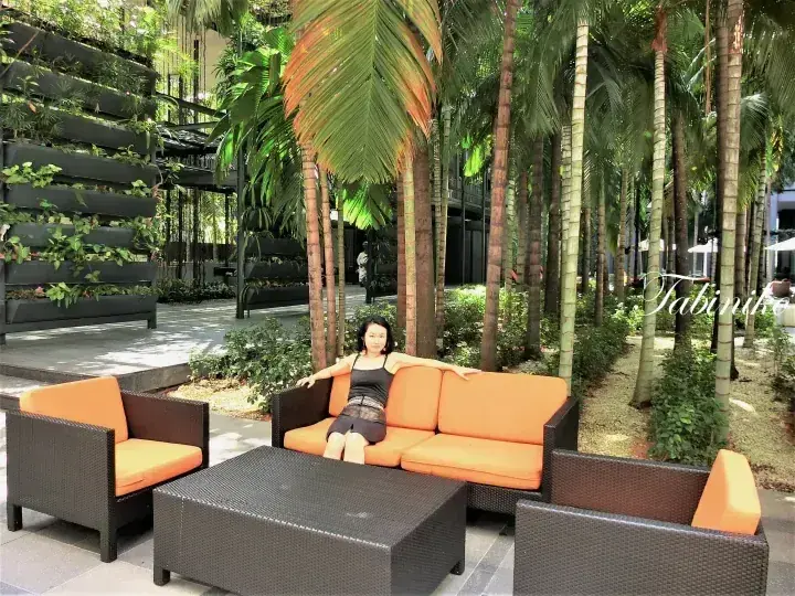 在現代物業的戶外，一位女士坐在被高大棕櫚樹和垂直綠化牆環繞的橙色沙發上放鬆。