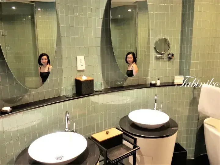 淺綠色磁磚浴室，配備兩個台上盆，鏡中映出一位女性。