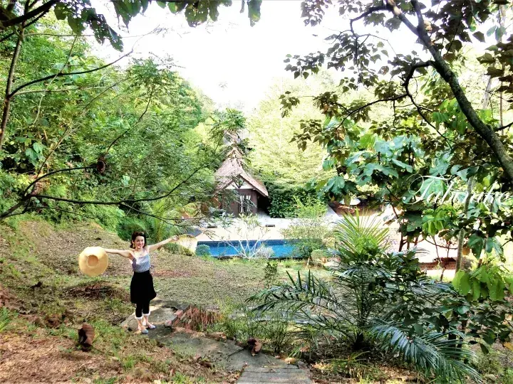 一位女士站在热带花园的小径上，背景是游泳池和木屋。