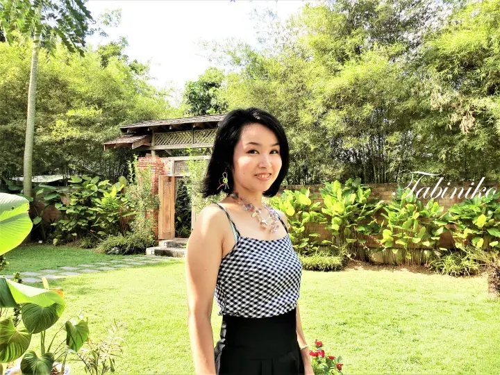 Woman in a checkered top standing in a lush garden with a wooden gazebo and bamboo trees.
