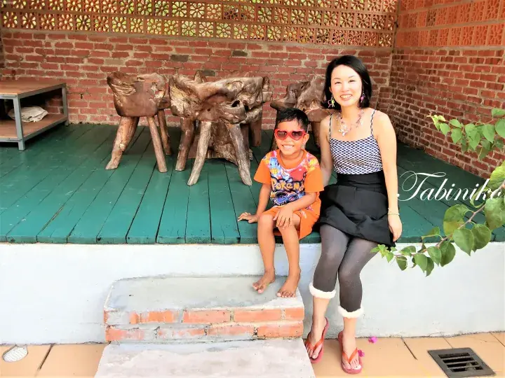 A woman and child pose on a green wooden deck with rustic wooden stools against a red brick wall.