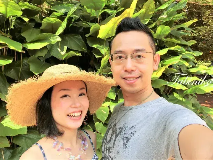 一对情侣在郁郁葱葱的热带绿植前自拍，画面右下角可见Tabiniko水印。