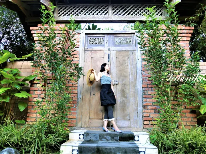 Woman holding a straw hat standing before a rustic wooden door framed by red brick and lush greenery.
