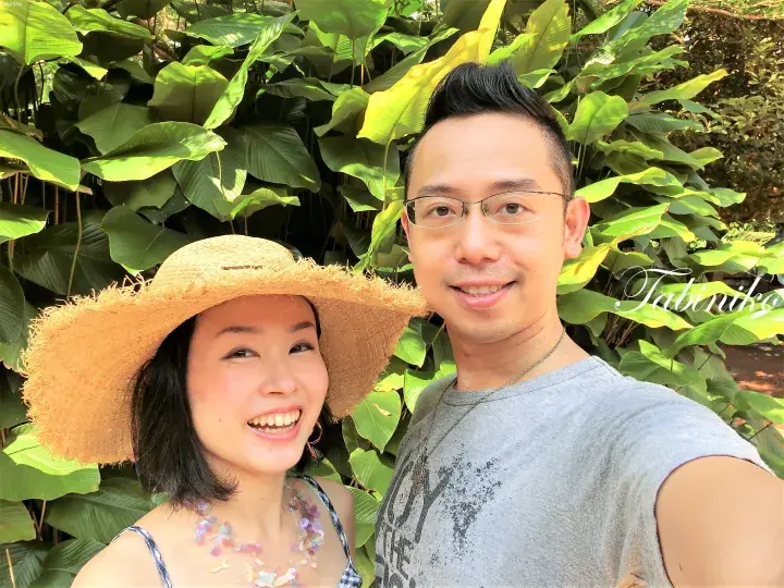 A smiling couple posing for a selfie in front of lush tropical greenery, with a Tabiniko watermark visible.