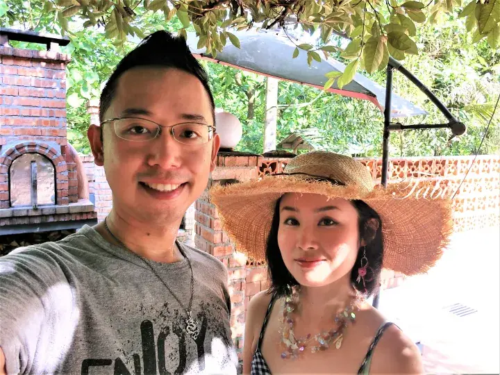 A smiling couple taking a selfie outdoors near a brick barbecue grill under leafy trees.