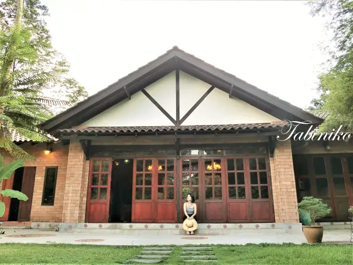 A woman sits on the steps of a heritage-style brick house with wooden doors and a gabled roof surrounded by lush greenery.
