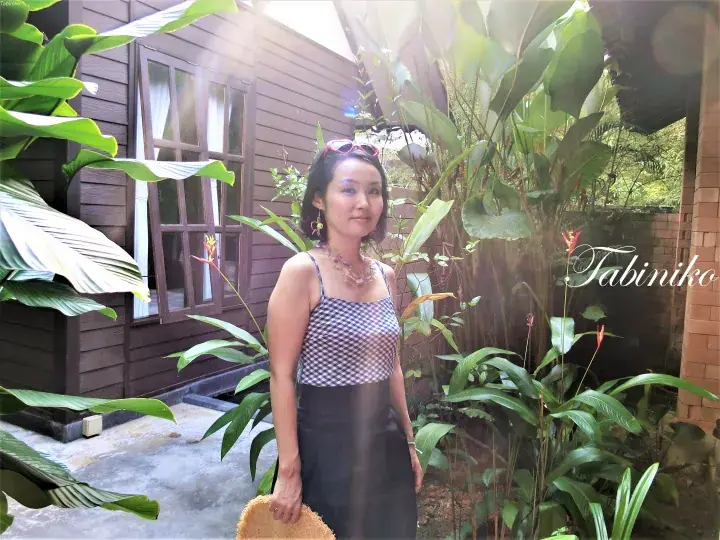 A woman stands in a lush garden outside a wooden cabin at Tabiniko, surrounded by tropical plants and sunlight.