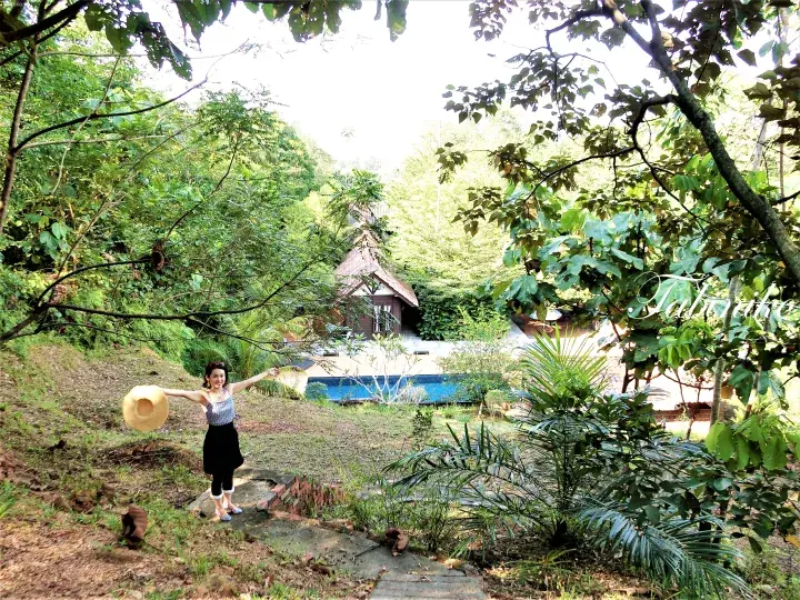 Woman holding a hat stands on a path in a lush tropical garden with a swimming pool and wooden cabin in the background.