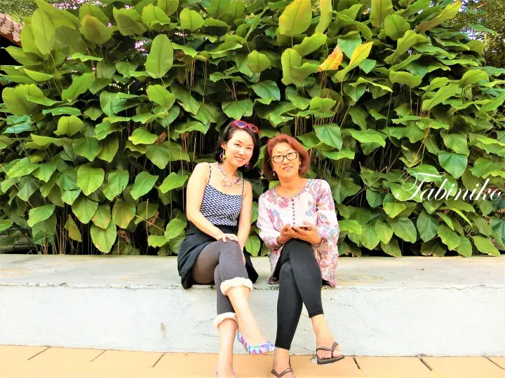 Two women sitting on a ledge in front of lush green foliage at Tabiniko, a property in Malaysia.