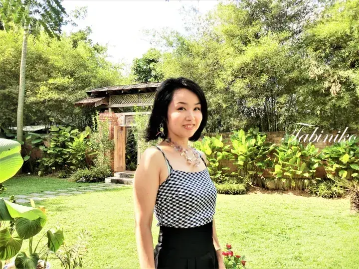 穿着格子上衣的女子站在绿意盎然的花园中，背景有木制凉亭和竹林。