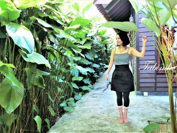 Woman posing on a garden walkway beside lush tropical plants near a wooden resort building.