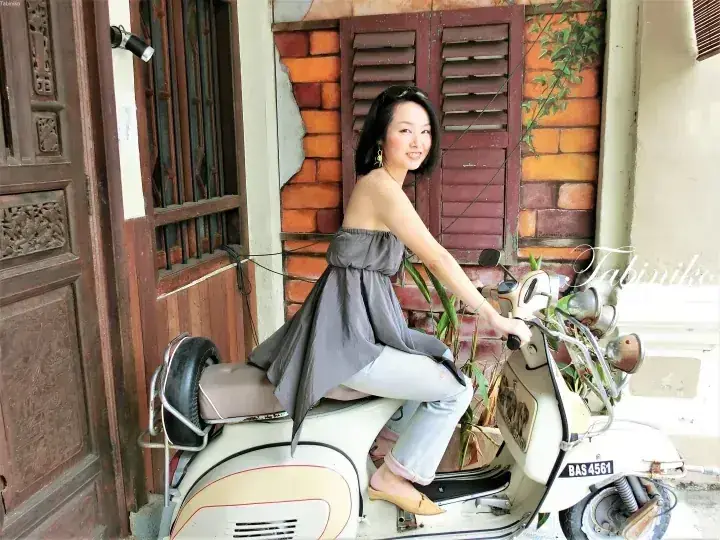 A woman sits on a vintage white scooter with license plate BAS 4561 in front of a heritage building with wooden shutters.