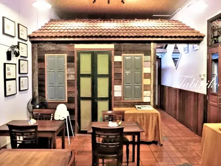 Interior featuring a traditional wooden house facade with green doors and windows, set within a dining area with tiled flooring.