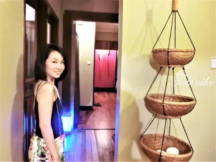 A smiling woman stands in a hallway next to a three-tiered hanging basket shelf in a property interior.