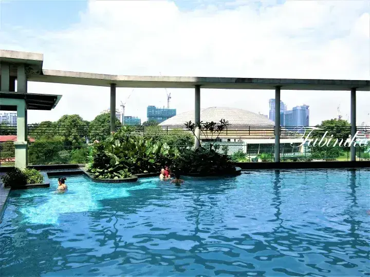 Swimmers enjoy a rooftop pool with lush greenery, overlooking the Kuala Lumpur skyline and a distinctive domed building.