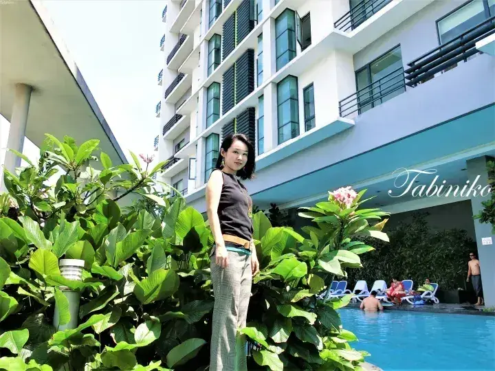 Woman standing by a poolside with lush greenery in front of a modern white hotel building.