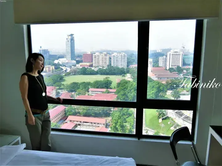 Woman standing by a large window overlooking a cityscape with green fields and buildings in Malaysia.