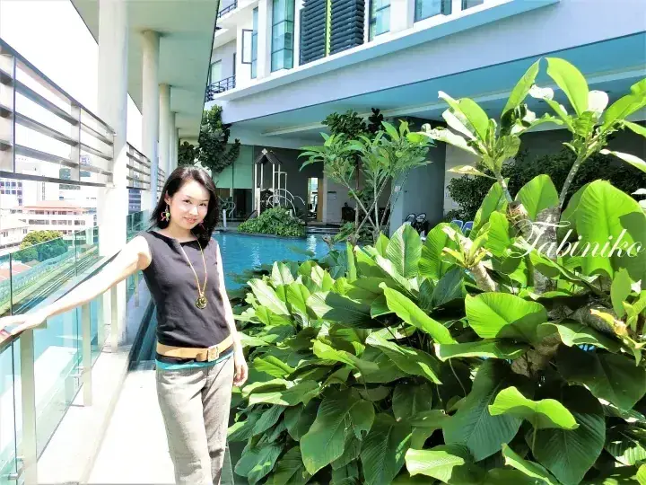 Woman standing on a balcony overlooking a swimming pool and lush tropical plants at a modern property.