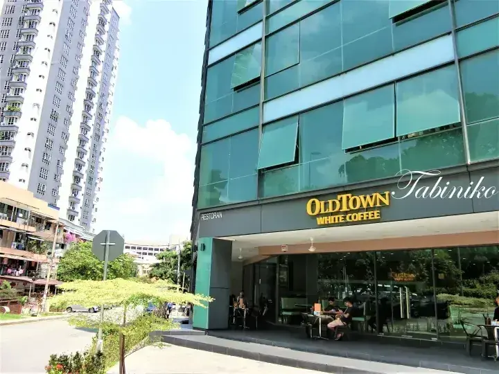 OldTown White Coffee storefront with glass facade and outdoor seating, located in a Malaysian urban area with high-rise residential buildings nearby.