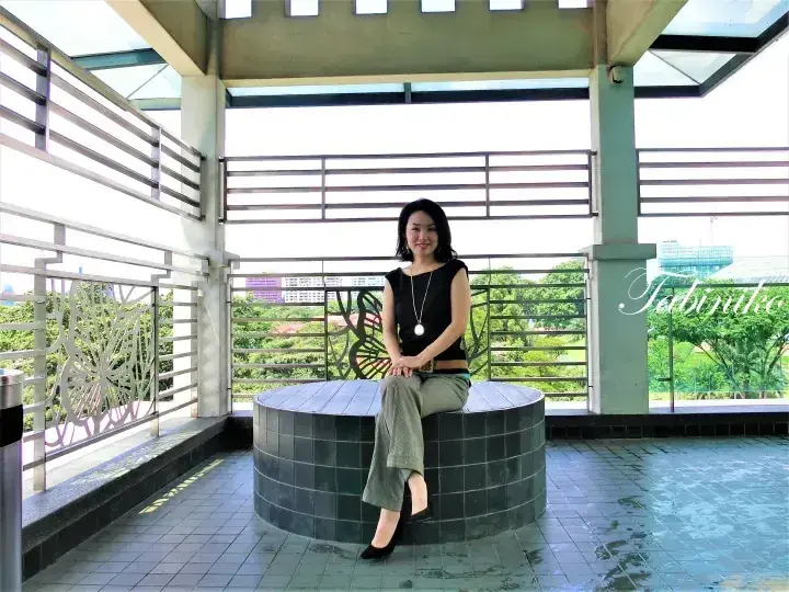 Woman sitting on a circular tiled bench at an outdoor pavilion with a view of greenery and city buildings.