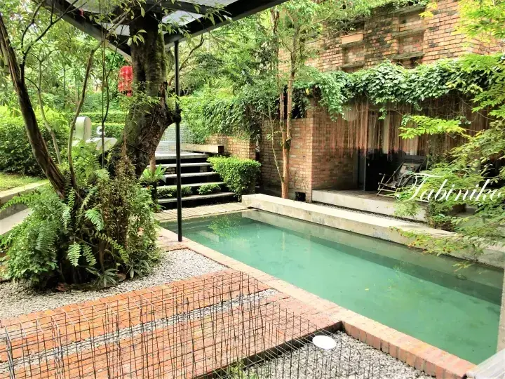 A tranquil private pool surrounded by lush greenery and a brick building, featuring a wooden deck and gravel garden area.