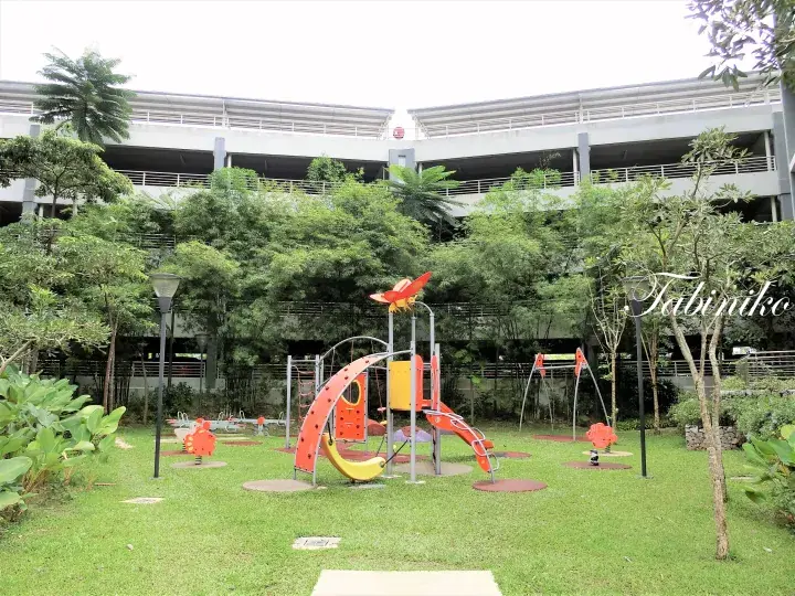 緑の芝生にカラフルな遊具があり、背景には木々と多層駐車棟が見える公園の風景。
