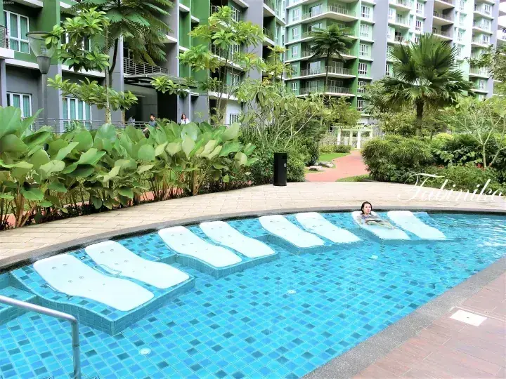 Swimmer relaxing in a pool with submerged lounge chairs at a modern apartment complex surrounded by lush greenery.