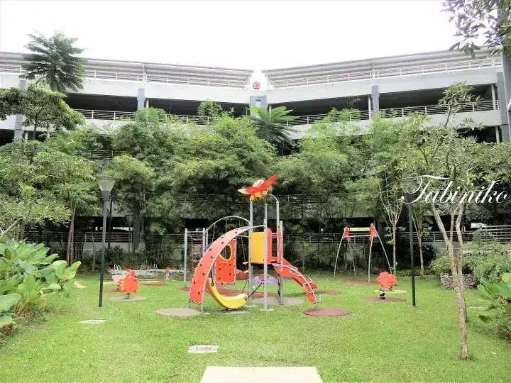 Colorful playground equipment on green lawn with trees and a multi-story car park building in the background.